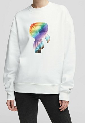 Osoba ubrana w białą bluzę z kolorowym nadrukiem kreskówkowej postaci w okularach przeciwsłonecznych i kucyku z przodu, w połączeniu z czarnymi spodniami.