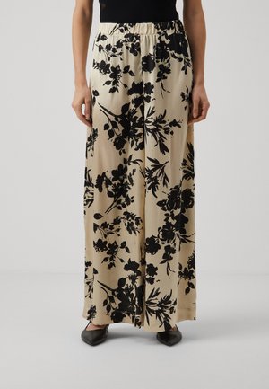 Beżowe spodnie z szerokimi nogawkami w czarny kwiatowy wzór, noszone z czarnymi butami na szpic i czarną bluzką, na jednolitym jasnym tle.