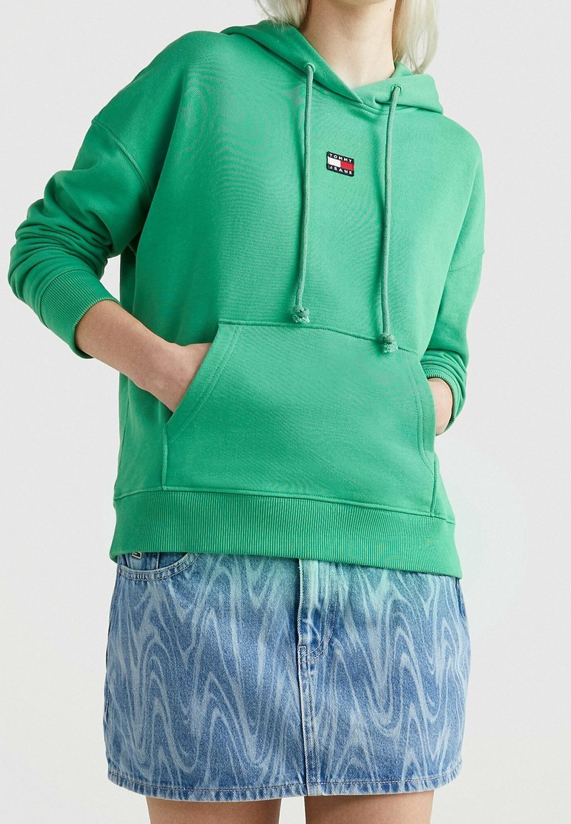 Sudadera verde con un bolsillo frontal, cordones y un pequeño parche con logo. Combinada con una falda de mezclilla azul clara y estampada.
