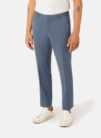 Unterer Teil einer Person, die hellblaue maßgeschneiderte Hosen, weiße Sneaker und ein weißes Hemd trägt und vor einem schlichten Hintergrund steht.