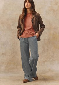 Chaqueta marrón a medida sobre una camiseta color óxido, combinada con vaqueros acampanados azul claro y zapatos de ante marrón. Fondo neutro.