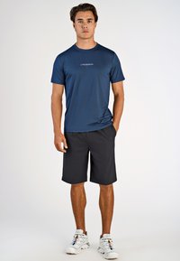 Mand iført en marineblå t-shirt med teksten "LINDBERGH", sorte shorts og hvide sneakers, stående med den ene hånd i lommen på hvid baggrund.