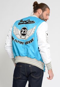 Lichtblauwe en witte bomberjack met geborduurde details, geribde manchetten en tailleband. Bevat patches: "TopGun," "23," en vleugels.