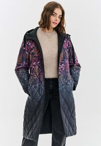 Žena v dlouhém prošívaném kabátu s fialovým květinovým vzorem, oblečená v béžovém svetru a černých džínách, stojící s rukama v kapsách.