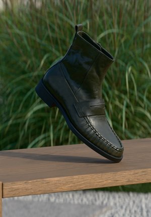 Black leather ankle boot with a low heel resting on a wooden surface with tall green grass in the background.