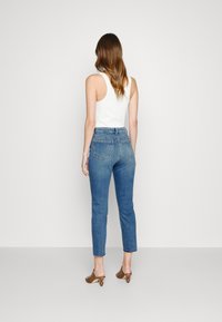Mujer con cabello largo vestida con una blusa sin mangas blanca, jeans cortos de cintura alta azul y mules de tacón marrones, de pie contra un fondo liso.