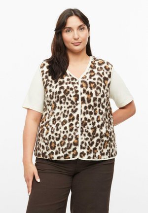 Woman with dark hair wearing a leopard print buttoned vest over a light short-sleeve shirt and dark pants, standing against a plain background.