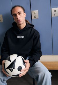 Čierna mikina s logom "JUVENTUS", modré džínsy, drží čierno-biely vzorovaný futbalový lopta, sedí pred skriňami.