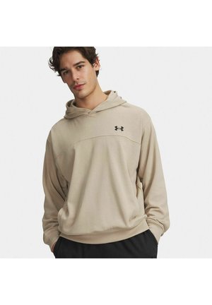 Giovane uomo che indossa una felpa con cappuccio beige Under Armour con le mani nelle tasche, in piedi contro uno sfondo chiaro e uniforme.
