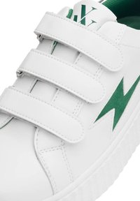 Sneaker blanc avec trois bandes Velcro, accent en éclair vert, semelle en caoutchouc texturée et doublure intérieure verte rembourrée.