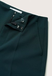 Pantalon vert foncé sur mesure avec fermeture à crochet et agrafe, bouton et zip, avec poche latérale visible.