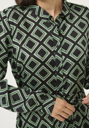 Vrouw die de tailleband van een zwart shirt met lange mouwen, een groen geometrisch vierkantpatroon en knopen aan de voorkant verstelt.