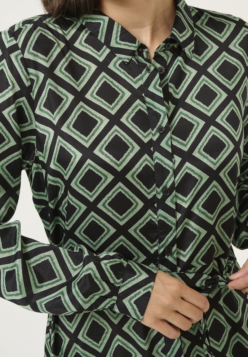 Femme ajustant la ceinture à la taille d'une chemise noire à manches longues avec motif géométrique carré vert et boutons sur le devant.