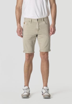 Hombre con pantalones cortos beige remangados, camiseta blanca y zapatillas blancas de pie contra un fondo liso.