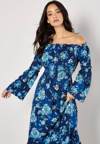 Marinblå blomstret maxi kjole med off-shoulder halsudskæring, lange klokkeærmer og smocket talje. Har blå og teal blomstermønstre over hele kjolen.