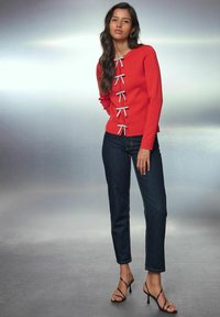 Vrouw in een rode blouse met lange mouwen en zilveren strikdetails, donkere spijkerbroek en zwarte hoge hakken, staand tegen een neutrale achtergrond.