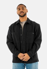 Chaqueta negra de botones con dos bolsillos frontales, fabricada en un tejido ligero, con cuello y un corte relajado, llevada sobre una camiseta de cuello redondo gris, combinada con jeans azules.