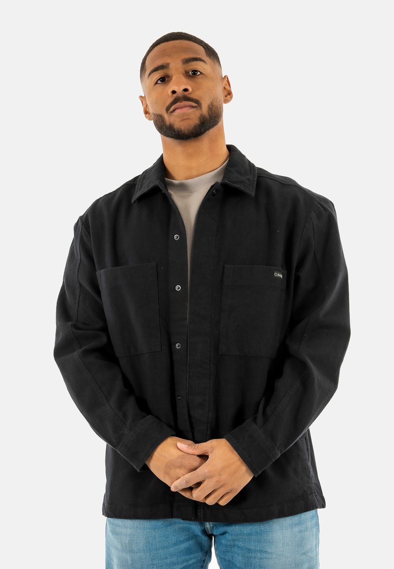 Chaqueta negra de botones con dos bolsillos frontales, fabricada en un tejido ligero, con cuello y un corte relajado, llevada sobre una camiseta de cuello redondo gris, combinada con jeans azules.