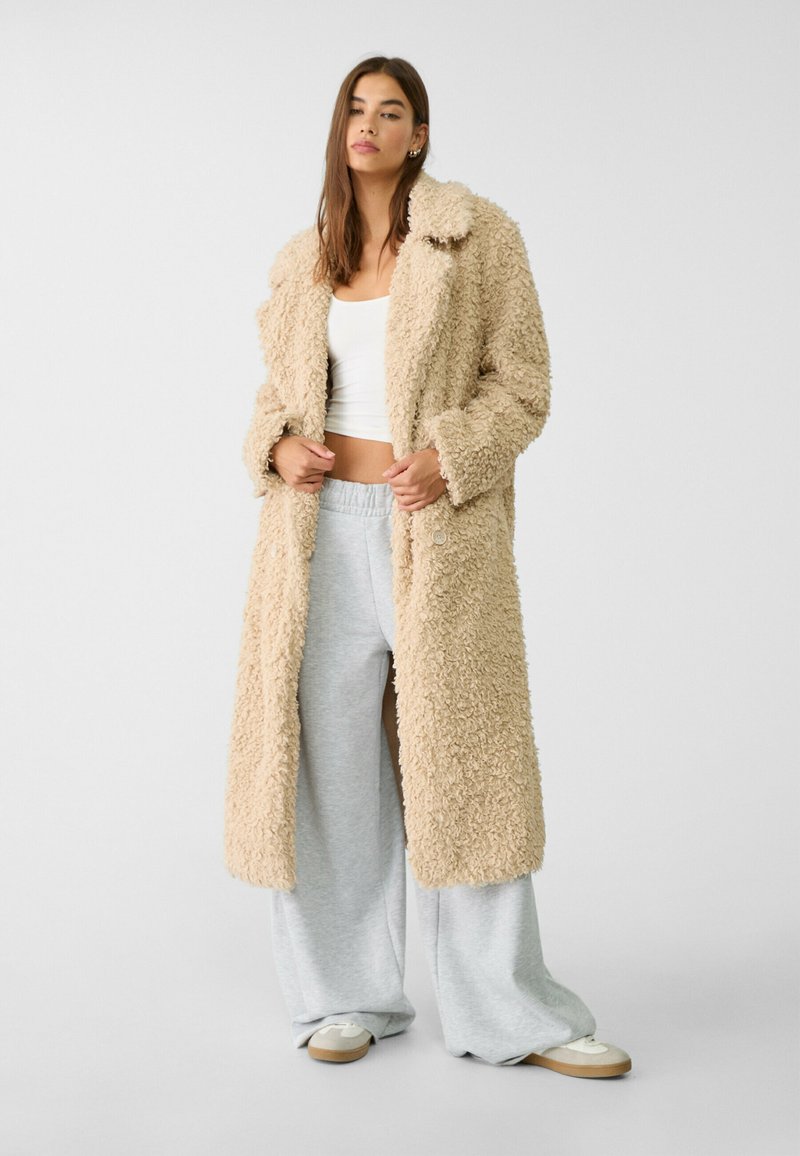 Beżowy, puszysty długi płaszcz o miękkiej fakturze, z dużymi klapami i dwoma guzikami z przodu, noszony na szare szerokie spodnie dresowe i jasne buty.