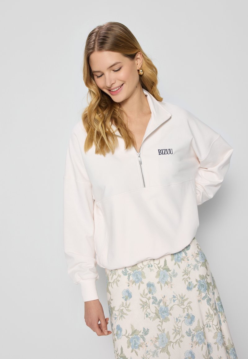 Lichtroze zip-up sweatshirt met een hoge kraag, met een witte panel en het "BIZUU" logo. Gecombineerd met een rok met een bloemenpatroon.