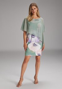 Abito floreale con sovrapposizione verde menta, caratterizzato da grandi fiori pastello. L'abito è aderente, con una texture liscia e maniche corte.