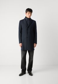 Casaco de marinha com um design ajustado, frente de um único botão, lapelas recortadas e dois bolsos laterais, combinado com jeans escuros e sapatos pretos.