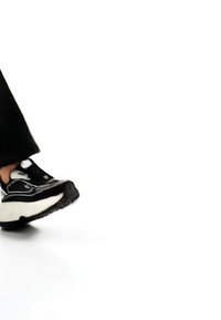 Stopa w czarno-białym trampku stawiająca krok do przodu na białym tle, ubrana w czarne spodnie.