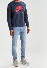 Námořnická mikina se červeným logem Nike Air, černými rukávy, v kombinaci se světle modrými džíny a šedými teniskami. Hladká struktura, volný střih.