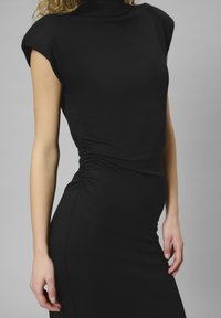 Rochie neagră cu guler înalt, mâneci scurte și detalii pliate pe lateral. Confectionată dintr-un material neted și elastic, urmează o siluetă body-con.