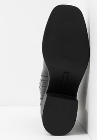 Suela de bota de cuero negro con tacón cuadrado, colocada verticalmente contra una pared blanca en la esquina, mostrando la parte inferior lisa y los detalles de costura en los lados.