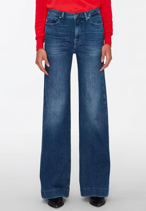 Kobieta ubrana w niebieskie dżinsy z wysokim stanem i szerokimi nogawkami, zestawione z czerwonym swetrem i czarnymi, spiczastymi butami.