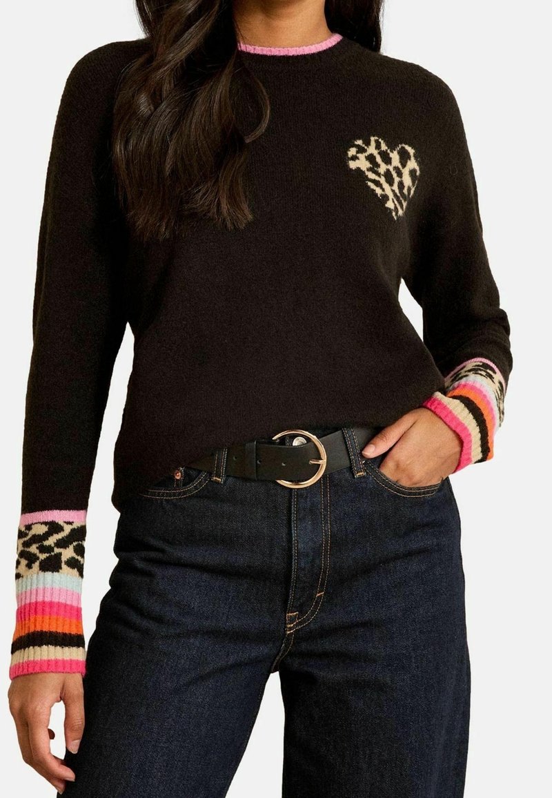 Femme portant un pull noir avec un cœur imprimé léopard sur la poitrine, des poignets rayés multicolores et un jean bleu foncé avec une ceinture noire.