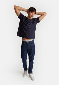 Granatowa krótka koszulka z napisem i ciemnoniebieskie dżinsy. Model nosi białe sneakersy, prezentując swobodny wygląd z luźnym krojem i stylem.