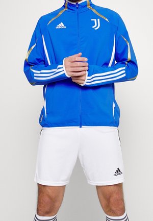 Hombre con chaqueta deportiva azul Adidas Juventus y pantalones cortos blancos Adidas, de pie con las manos suavemente entrelazadas a la altura de la cintura.