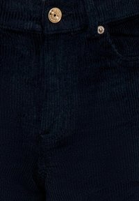 Kratki prikaz tamnoplave kord pantalone s zlatnim dugmetom i malim metalnim zakovicom blizu džepa.