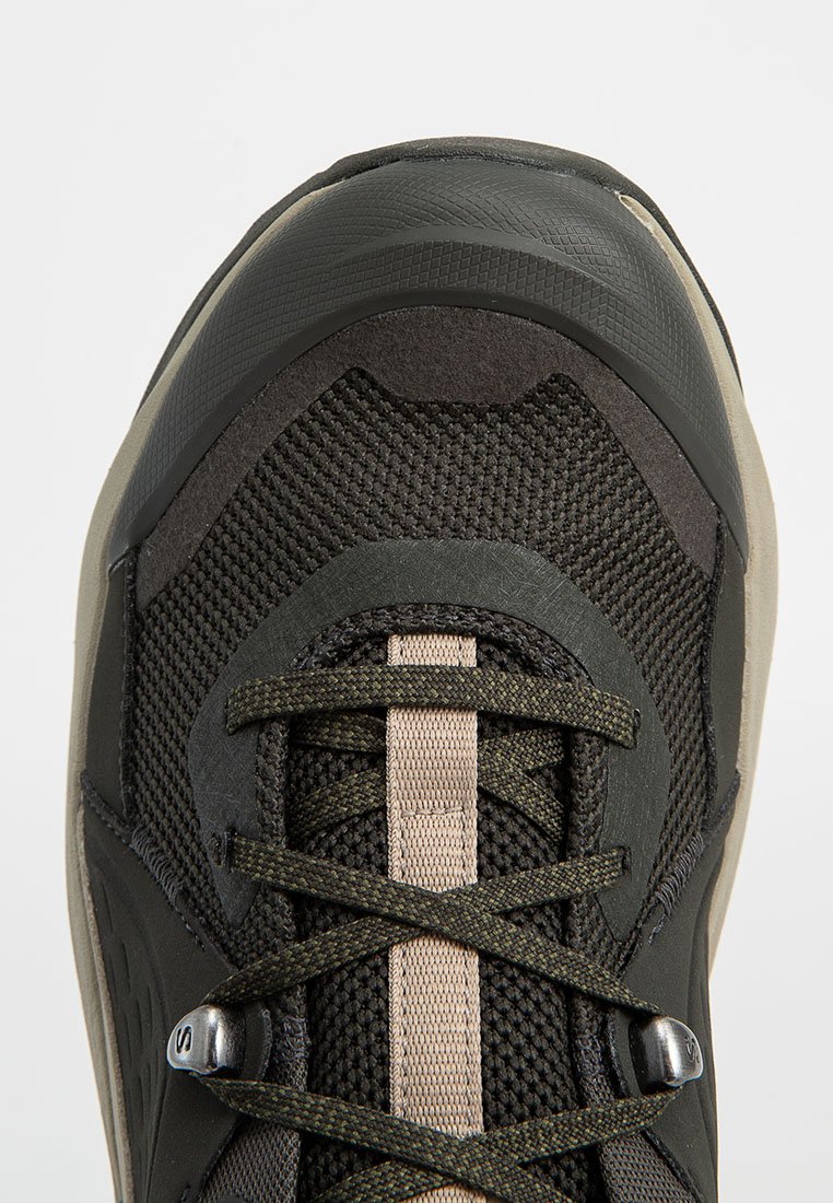 Zapato de senderismo para hombre con parte superior de malla negra y oliva, cubrepunta de goma, acentos beige y ojales reforzados con cordones texturizados.