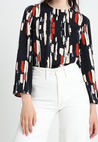 Långärmad blus i marinblått med abstrakt penseldragmönster i röd, krämfärgad och svart. Slätt tyg med rund halsringning. Vita högmidjade byxor.