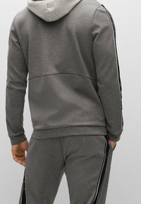Uomo vestito con felpa grigia con cappuccio e pantaloni della tuta abbinati con strisce laterali nere e bianche, rivolto di spalle, mani rilassate lungo i fianchi.