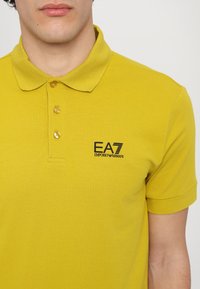 Sárga pamut póló, galléros nyakkal, három gombbal, és fekete EA7 logóval a bal mellrészen.