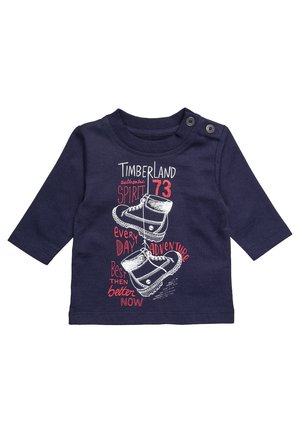 Marineblauw shirt met lange mouwen met een afbeelding van laarzen en de tekst: "Timberland authentic spirit 73," "elke dag avontuur," en "beter nu."