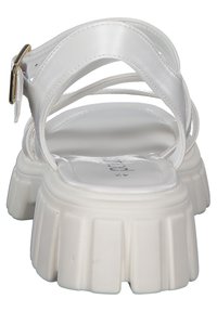 Valkoinen sandaali, jossa on paksu pohja, ristikkäinen strapisuunnittelu ja metallinen solki yksityiskohtana nilkassa. Silkkinen nahkamateriaali.