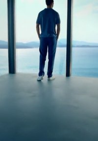 Lässiges Outfit mit einem blauen Hemd und passenden Hosen, stehend auf einem glatten Boden neben einer Glaswand mit Blick auf Wasser und Berge.