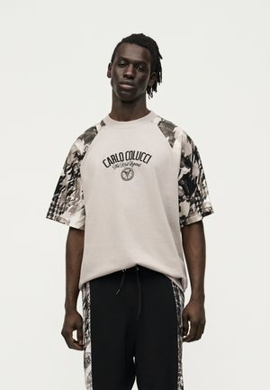 Νεαρός άντρας με σκουρόχρωμο δέρμα και πλεξούδες, φορεί μια μπεζ και μαύρη μπλούζα Carlo Colucci με τύπωμα και αντίστοιχο μαύρο παντελόνι με πλευρικές ρίγες.