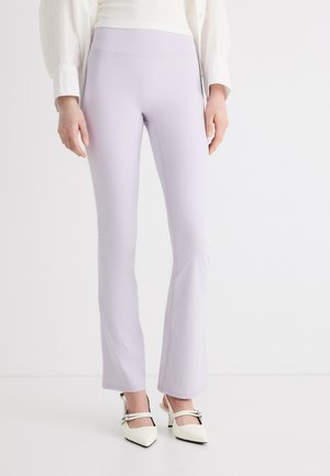 Naine, kes kannab helelilla tooniga kitsaid pükse, mille sääred on veidi lõhenevad ja valgeid rihmikute ja kontsadega kingi.