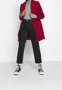 Rød lang frakke over en grå ribbet turtleneck, parret med højtaljede sorte jeans og sorte sneakers med hvide snørebånd og detaljer.