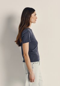 Young woman with long brown hair wearing a dark grey t-shirt and light blue jeans, standing in profile against a plain beige wall.