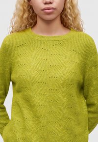 Jeune personne aux cheveux blonds bouclés portant un pull tricoté texturé vert vif avec un col rond.