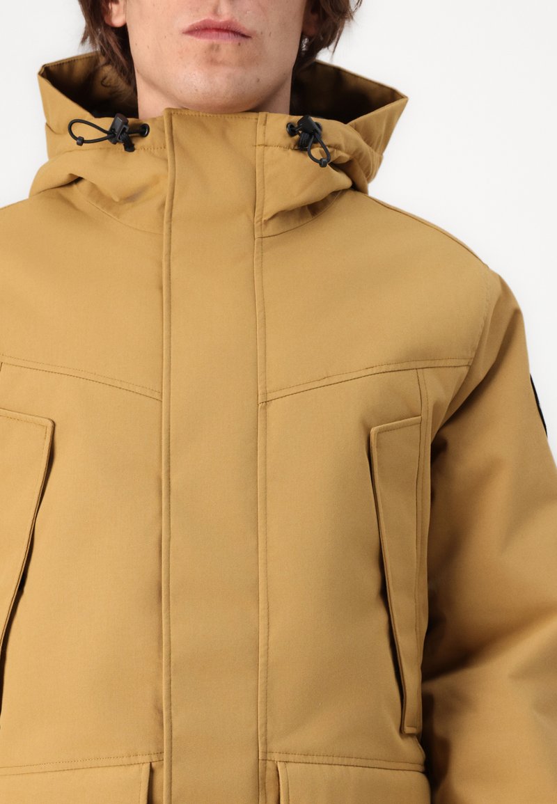 Chaqueta aislante color crema con cuello alto, capucha ajustable y dos bolsillos frontales, de textura suave y con detalles de costura mínimos.