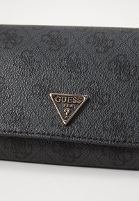 Fekete textúrájú pénztárca kiemelkedő monogram mintával és arany háromszög Guess logóval az elején. Biztonságos és kompakt kialakítás.