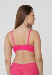 Růžový krajkový bralet s nastavitelnými ramínky a zapínáním na háček, zdobený květinovými vzory a vlnitým okrajem na zadní straně.